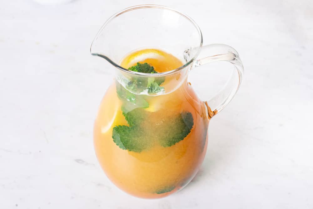 peach lemonade in a glass pitcher with peach slices and mint floating in it.