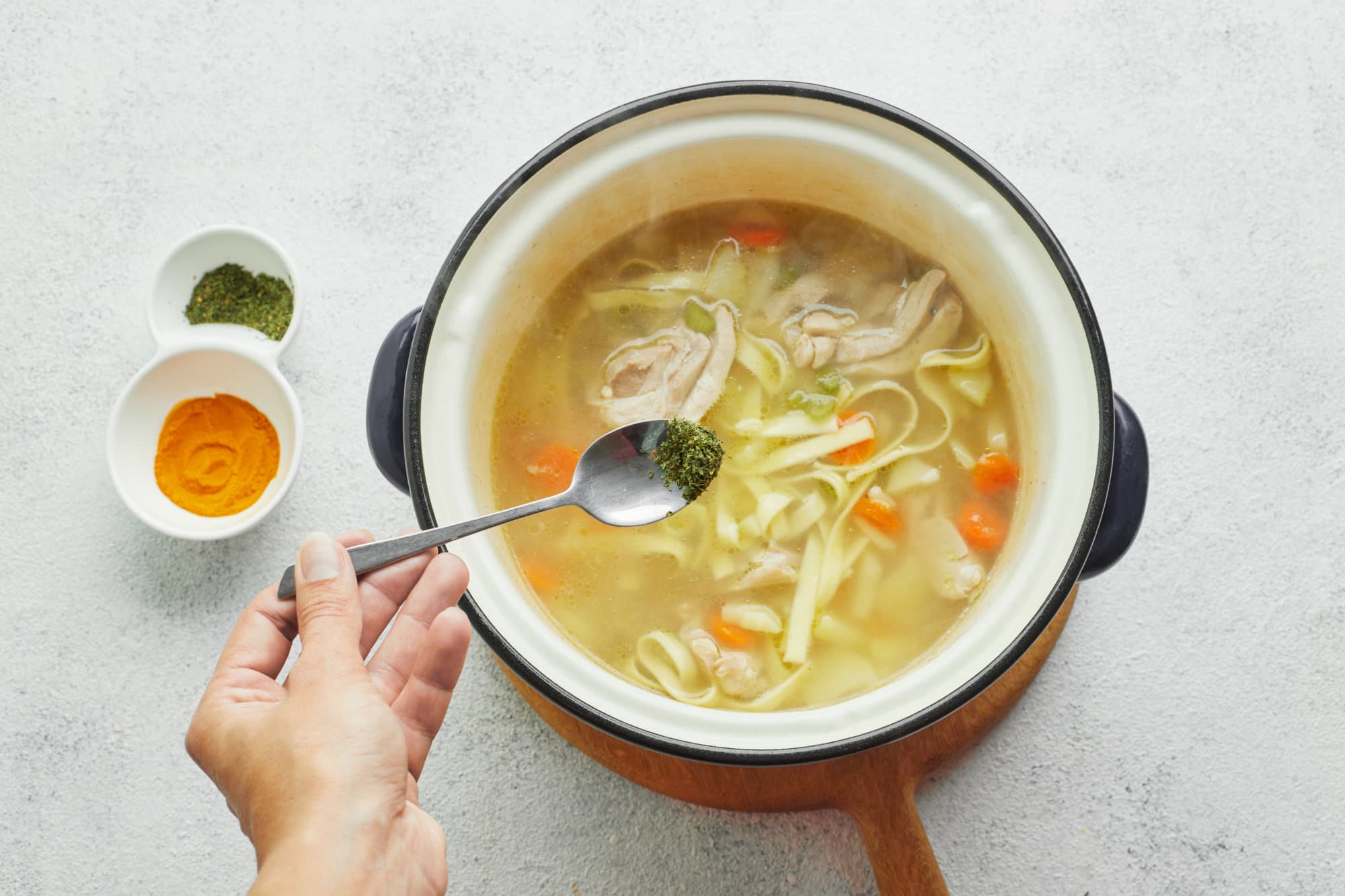 chicken-soup-in-a-pot-on-a-wooden-baord-with-spices-being-added-in-with-a-spoon-with-two-bowls-of-spices-on-the-side