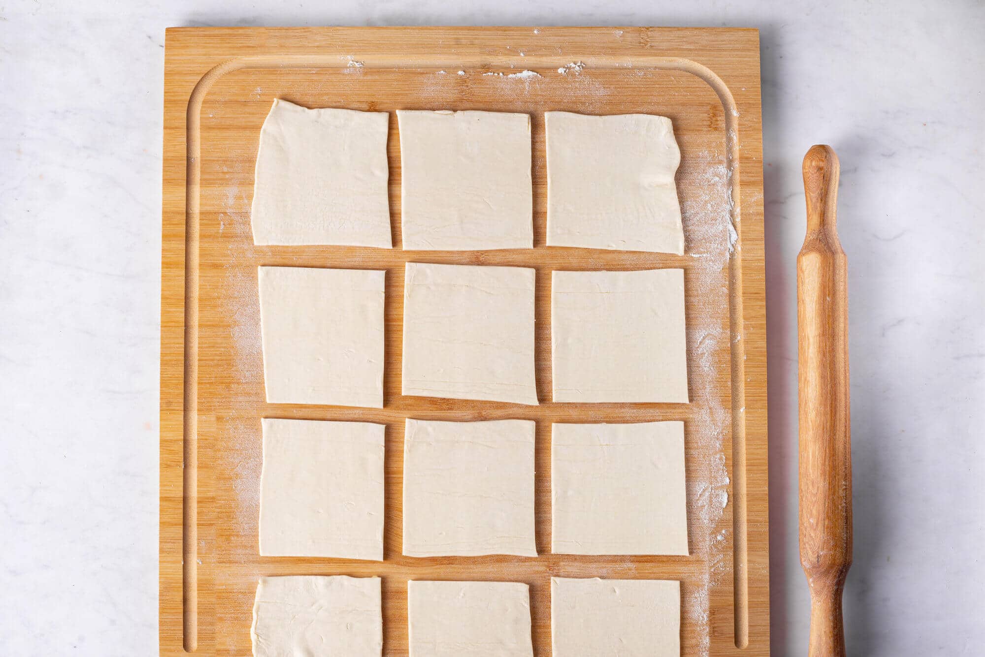 Puff pastry cut into squares on a wooden cutting board with a rolling pin on the side.