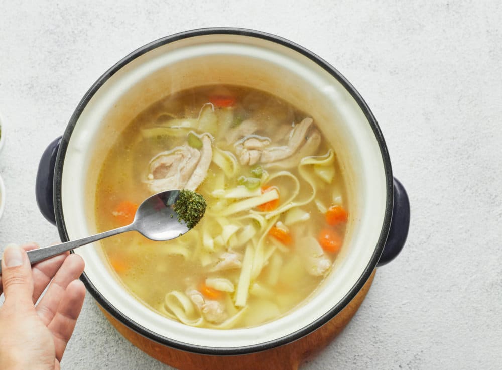 chicken-soup-in-a-pot-on-a-wooden-baord-with-spices-being-added-in-with-a-spoon-with-two-bowls-of-spices-on-the-side