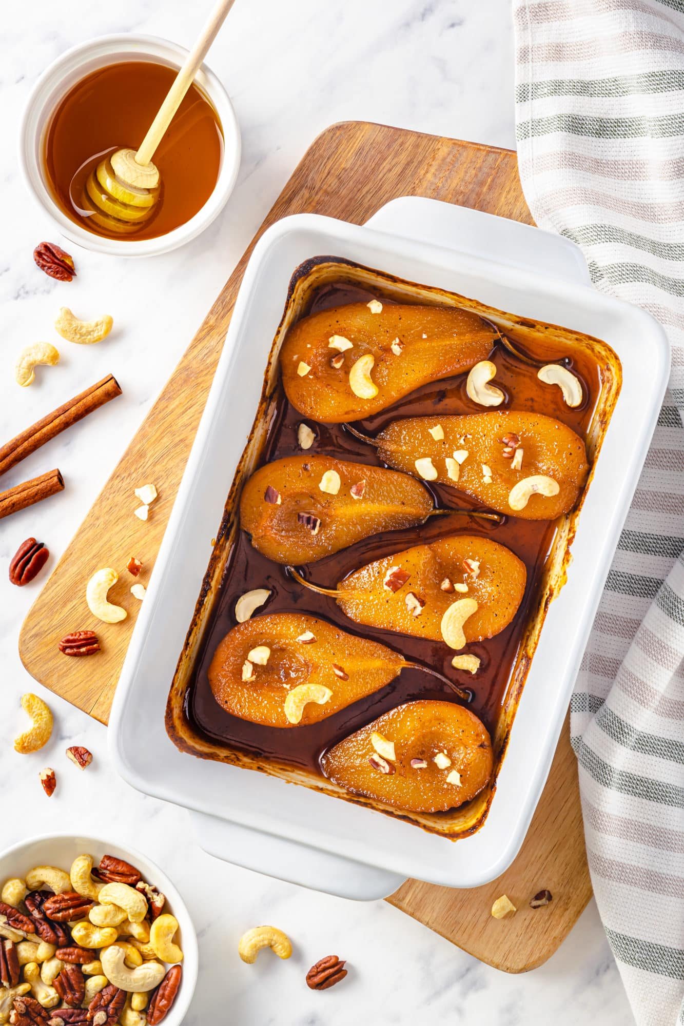 roasted pears in a baking tray on a wooden board with nuts on the side and a grey towel with a bowl of honey