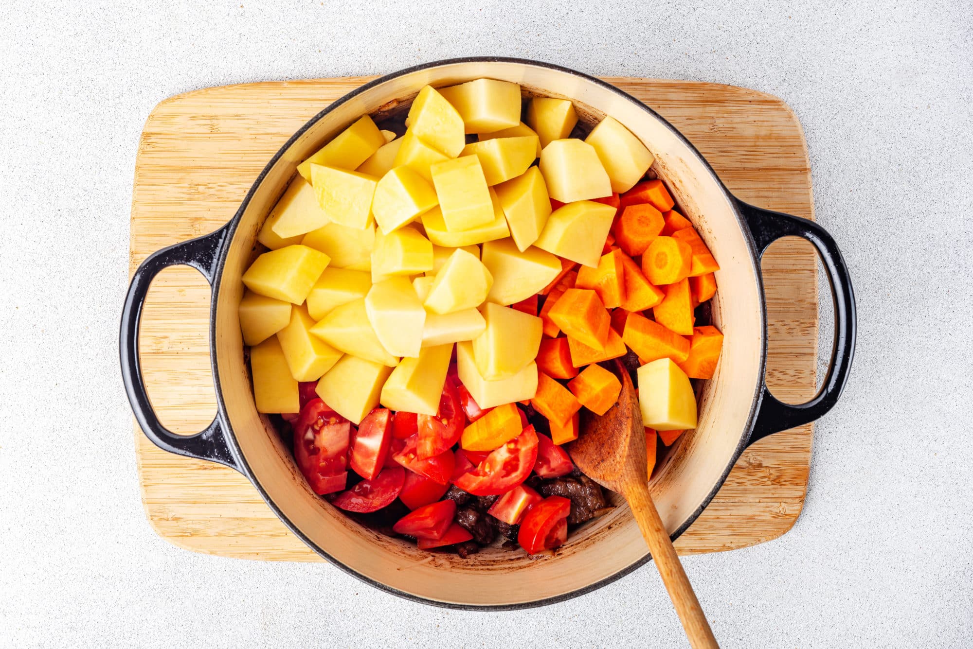 A wooden spoon carrots tomatoes potatoes and meat in a large pot on a wooden board.