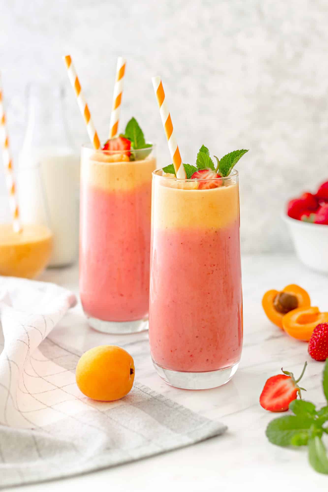 Two clear glasses filled with apricot and strawberry smoothie with striped straws topped with mint and fruit.