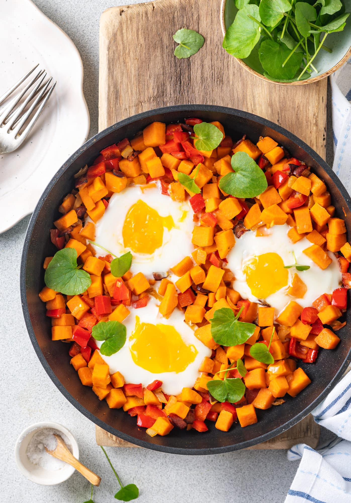 Sweet potato and eggs in a skillet with a sprinkle of micro greens on a wooden board and forks and a small bowl of salt on the side. 