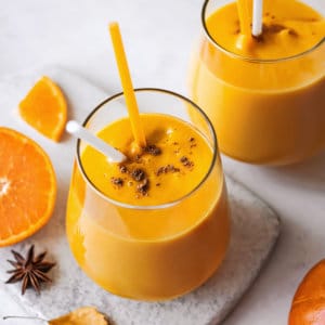 Pumpkin smoothie in a glass cup with a straw and topped with pumpkin seeds and spices on a board.