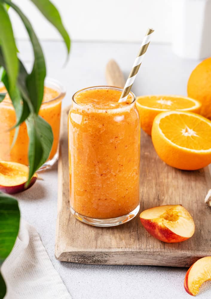 apricot smoothie in a glass cup with a straw and more halved oranges on the side.