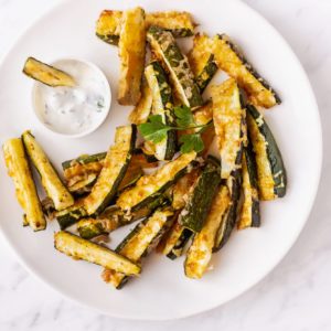 Roasted zucchini bites topped with cheese on a white plate with a bowl of dip.