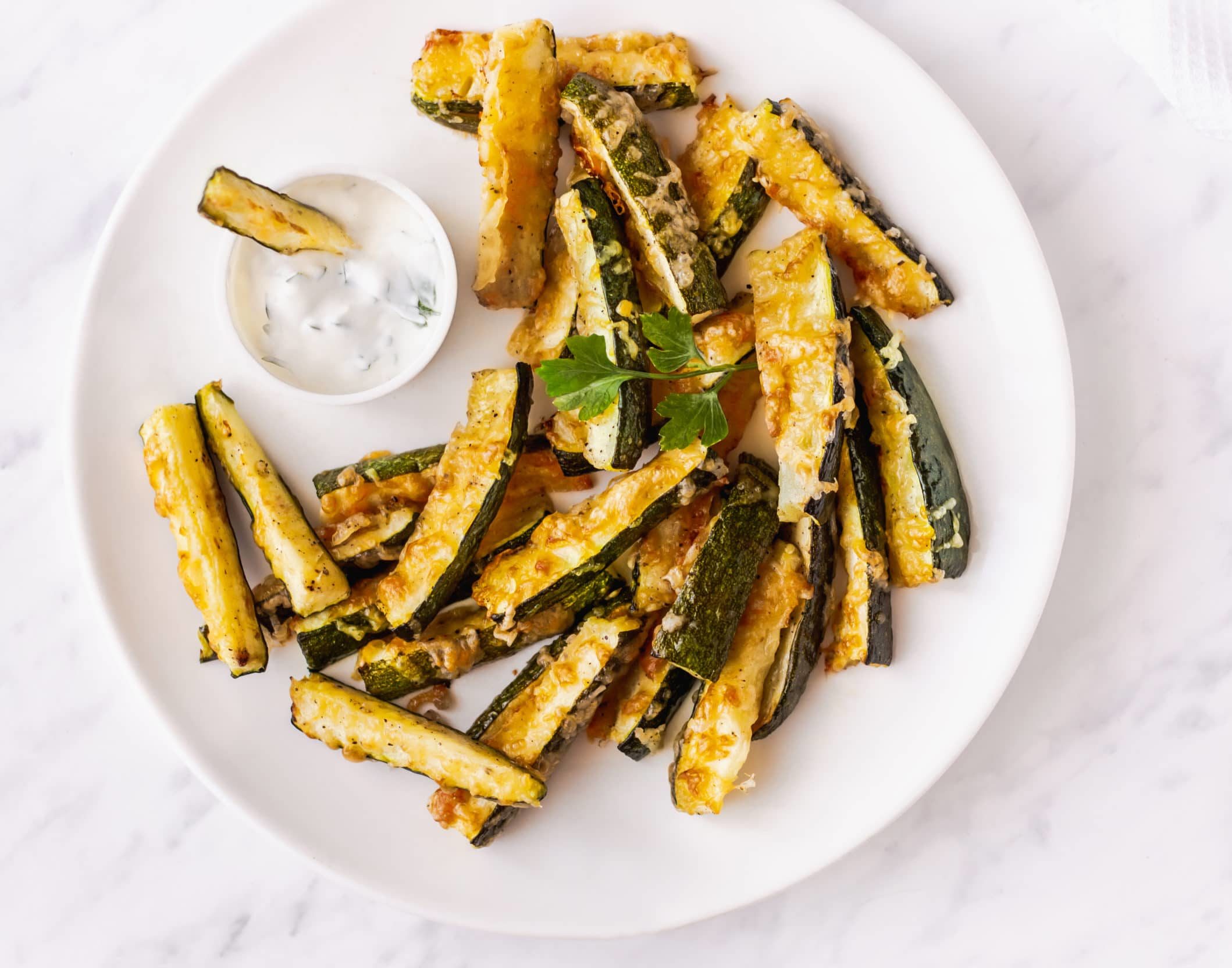 Roasted zucchini bites topped with cheese on a white plate with a bowl of dip.