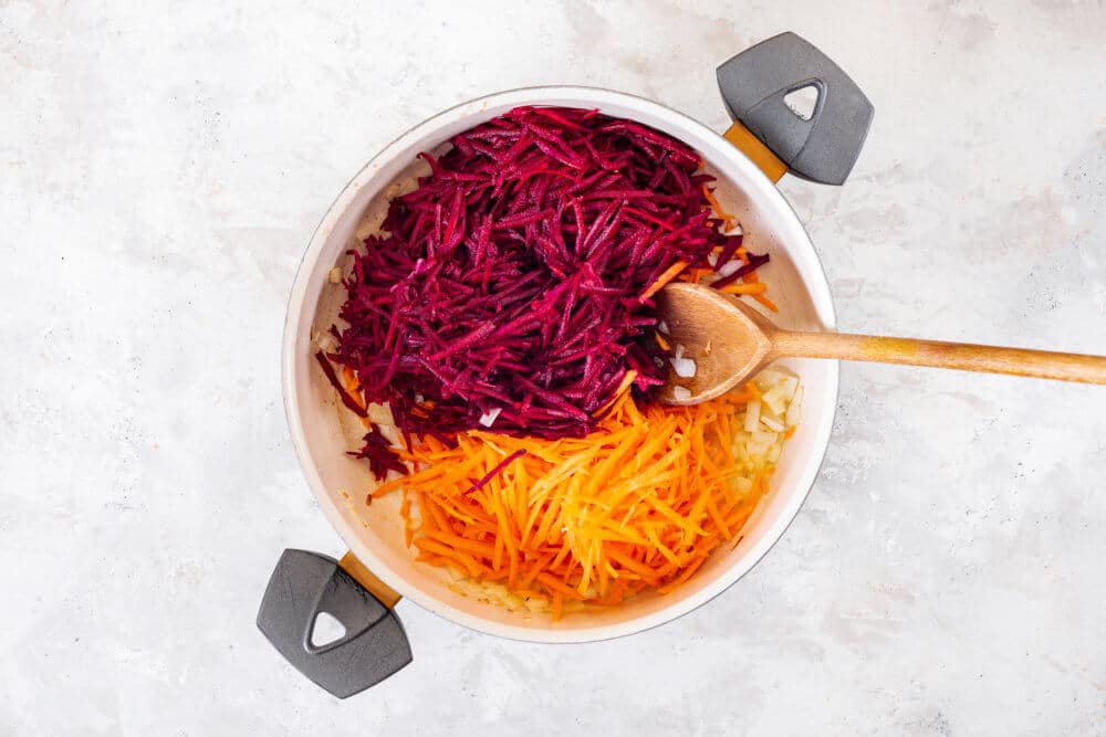 Carrots, beets, onion sauteing in a pot and mixing with a wooden spoon.