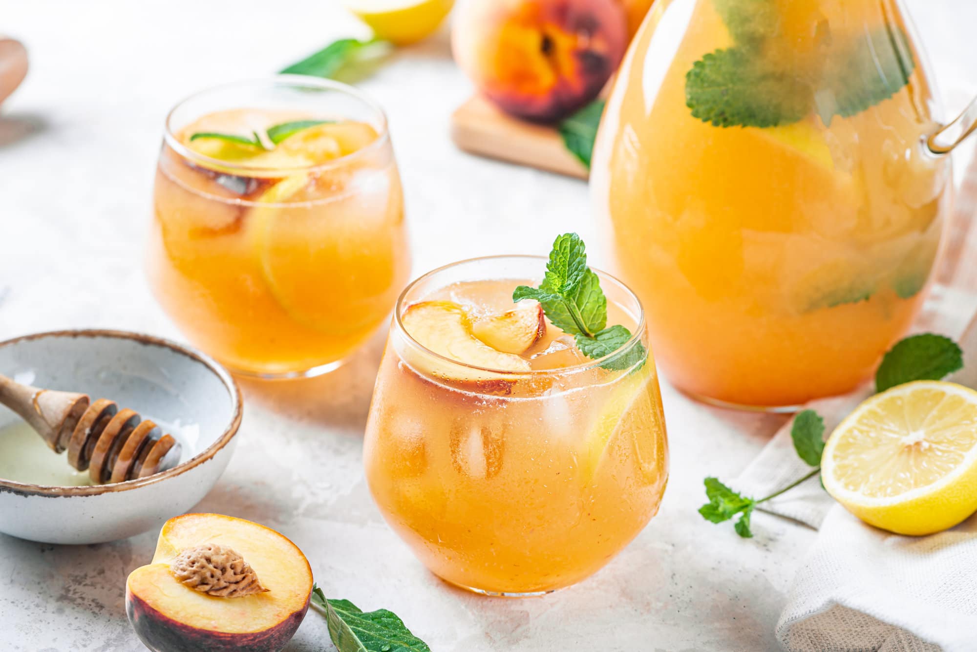 Peach lemonade in a glass cup with a pitcher on the side and fresh peaches around.