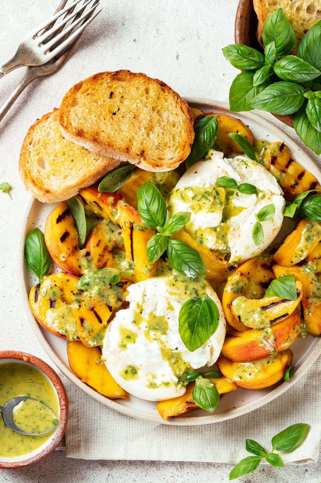 a plate with peaches burrata and toasted bread slices with basil and dressing on top and silver forks and more basil on the side.