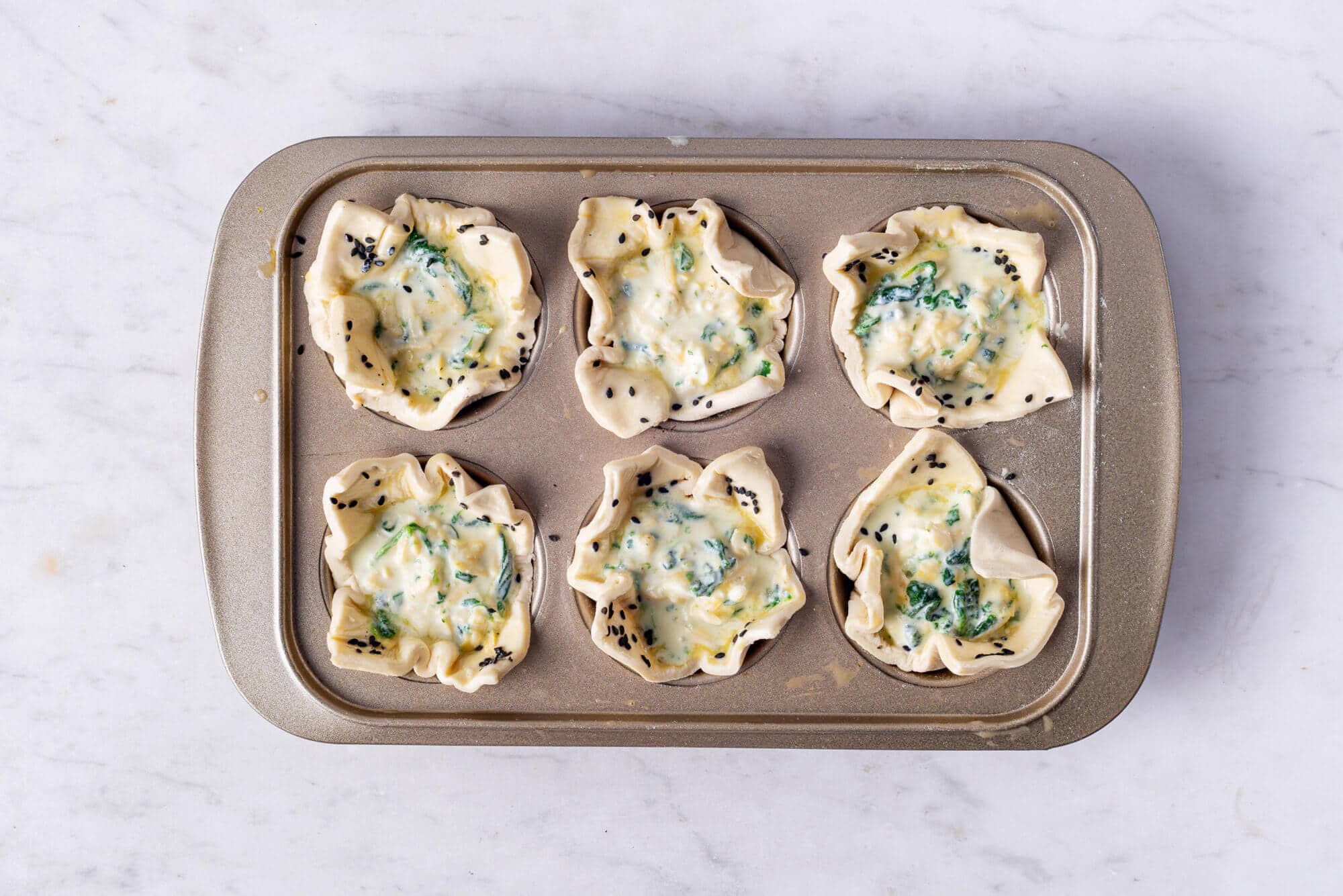 Raw spinach puffs in a muffin tray topped with black sesame seeds. 