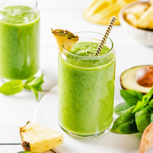 two glasses filled with a citrus green smoothie with a straw and pineapple wedge with ingredients on the side.