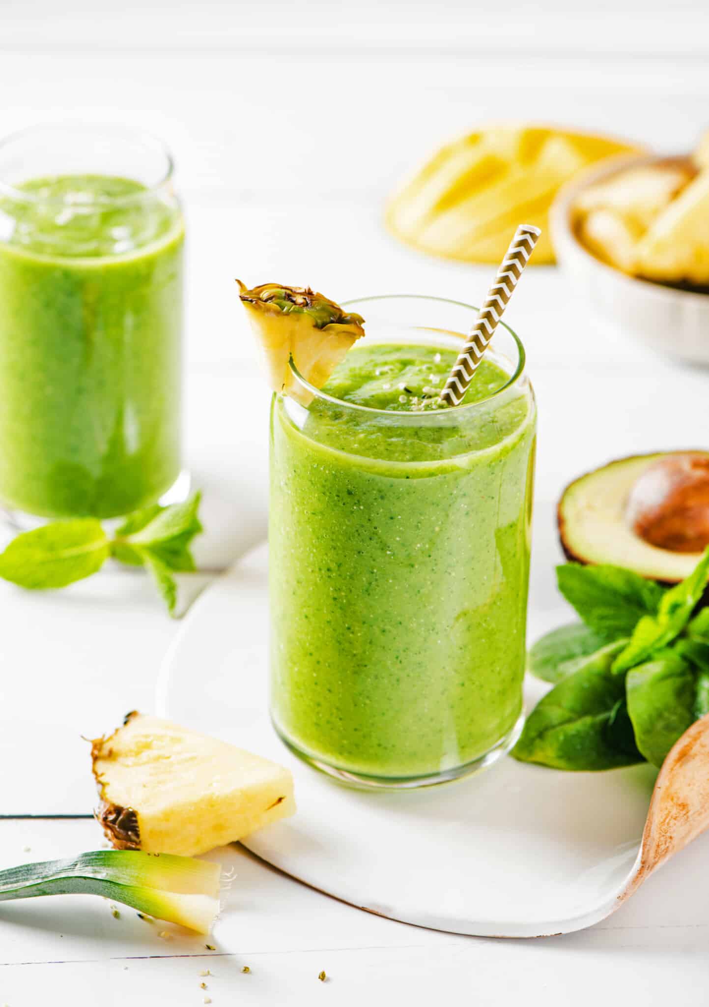 two glasses filled with a citrus green smoothie with a straw and pineapple wedge with ingredients on the side.