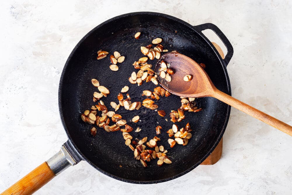 almonds toasting in a skillet with a wooden spoon on a wooden board.