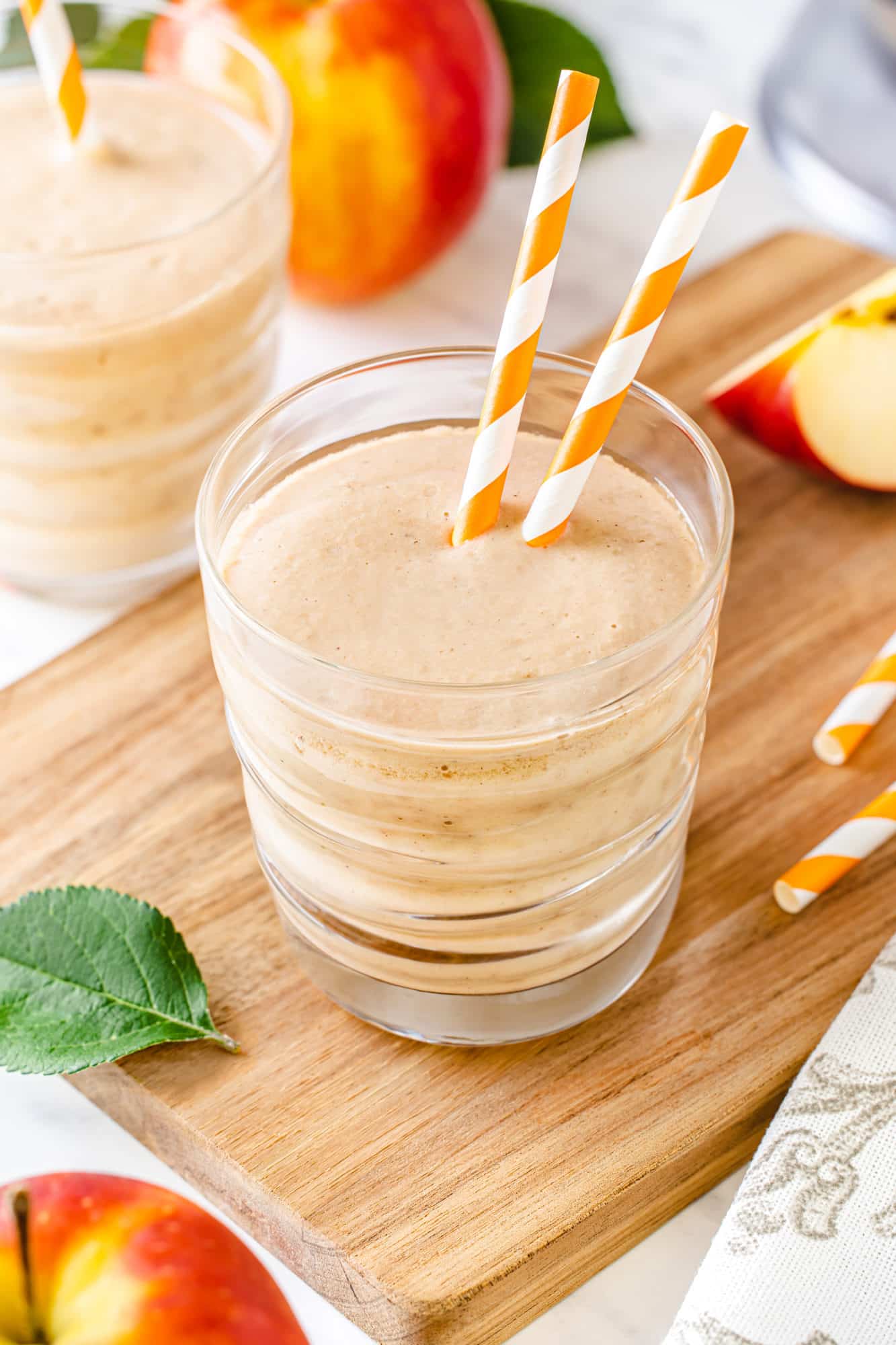 one glass with apple smoothie and two straws in the smoothie on a wooden cutting board.