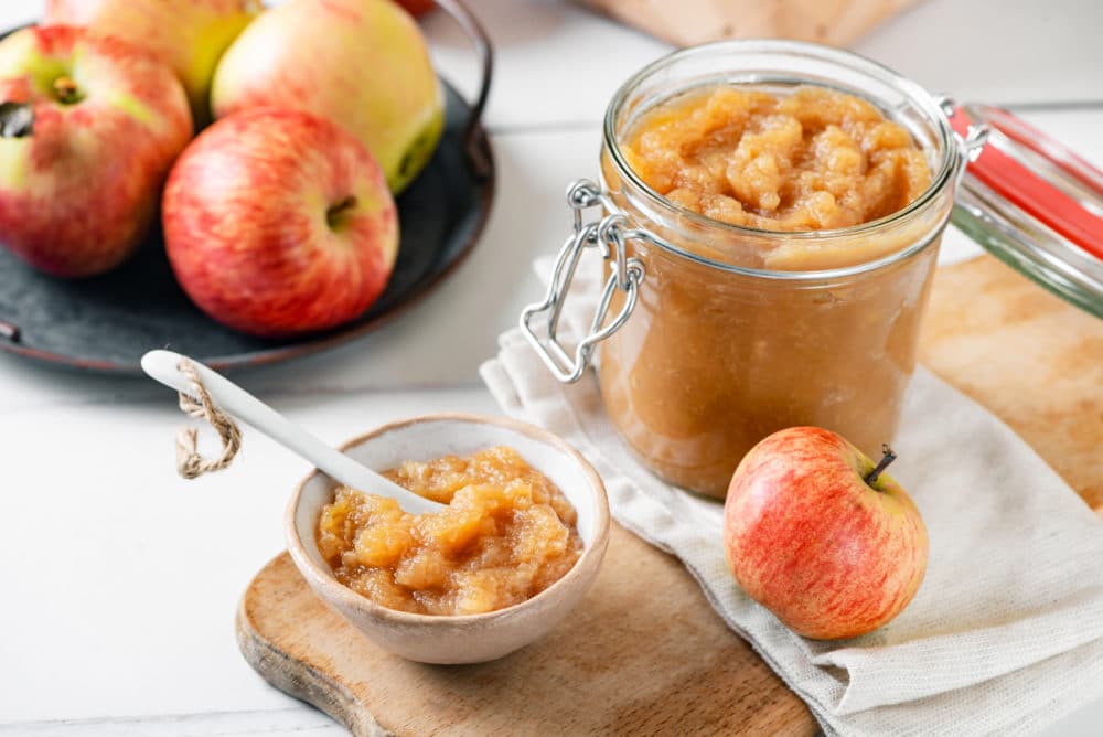 A glass jar and a white bowl of apple jam with a spoon in the bowl and apples all around.