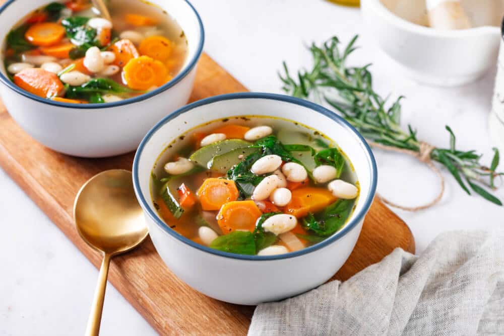 tuscan soup in a white bowl with a blue rim on a wooden board with a gold spoon on the side.