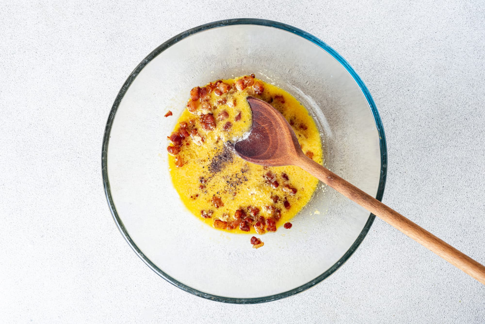 Beaten eggs and pancetta in a glass bowl with a wooden spoon, mixing.