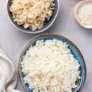 Three bowls one filled with cooked white rice one filled with cooked brown rice and one filled with uncooked white rice plus a towel on the side.