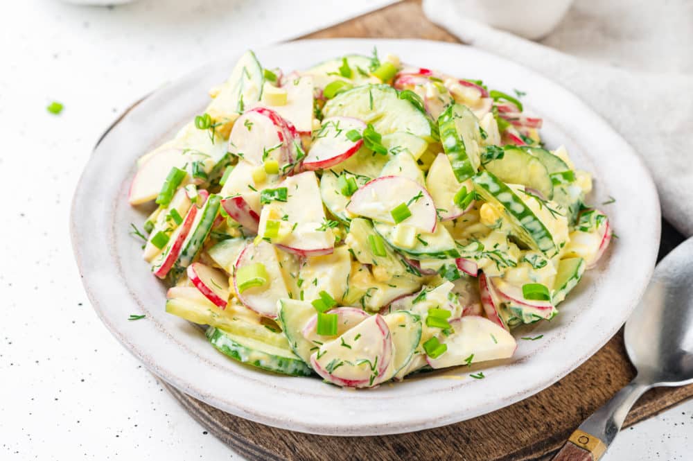 wooden-board-under-white-plate-with-radish-cucumber-salad-with-dill-and-green-onion-and-dressing-and-a-silver-spoon