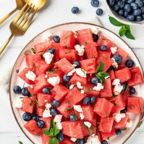 Feta and watermelon salad with mint on a white plate with a brown rim and a gold fork on the side.