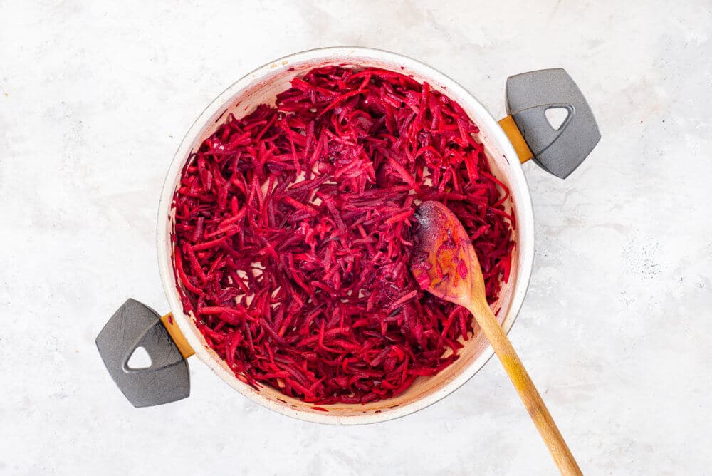 Grated beets added to a pan with a wooden spoon for mixing.