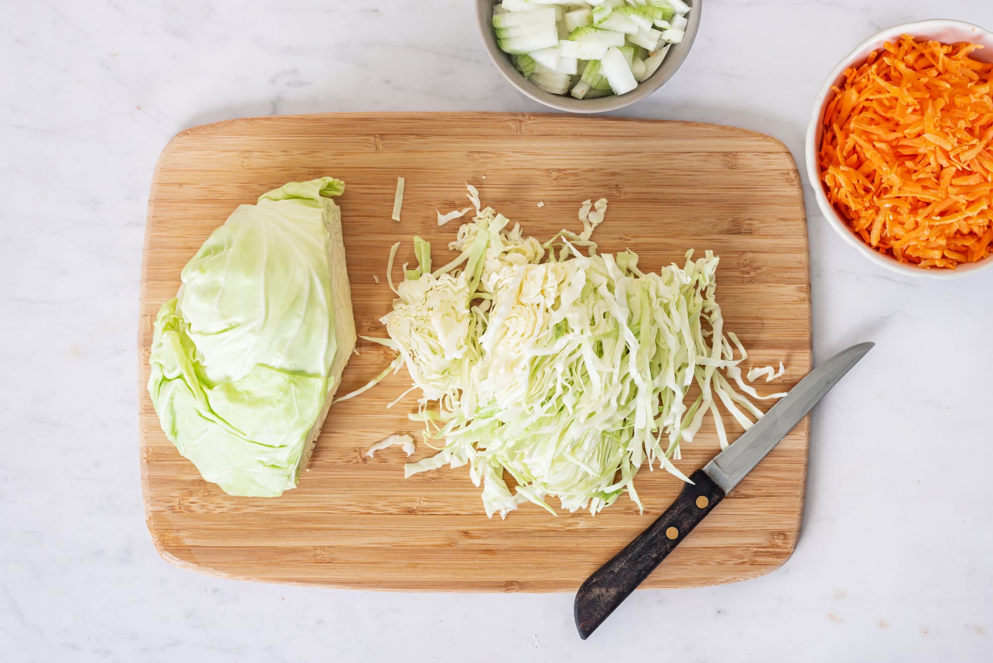 cabbage-shredded-on-a-wooden-board-with-a-knife-on-the-side-and-a-bowl-of-shredded-carrots-and-celery-in-a-bowl-on-the-sides