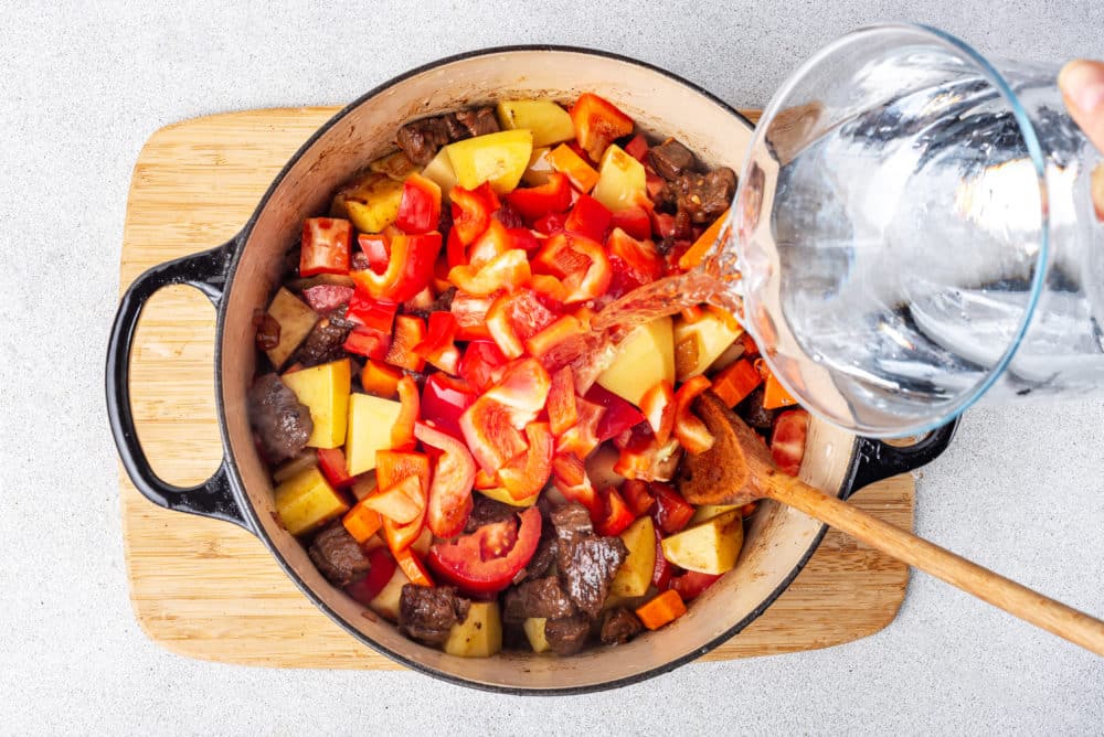 Soup ingredients in a large pot with water being poured into the pot from a glass pitcher.