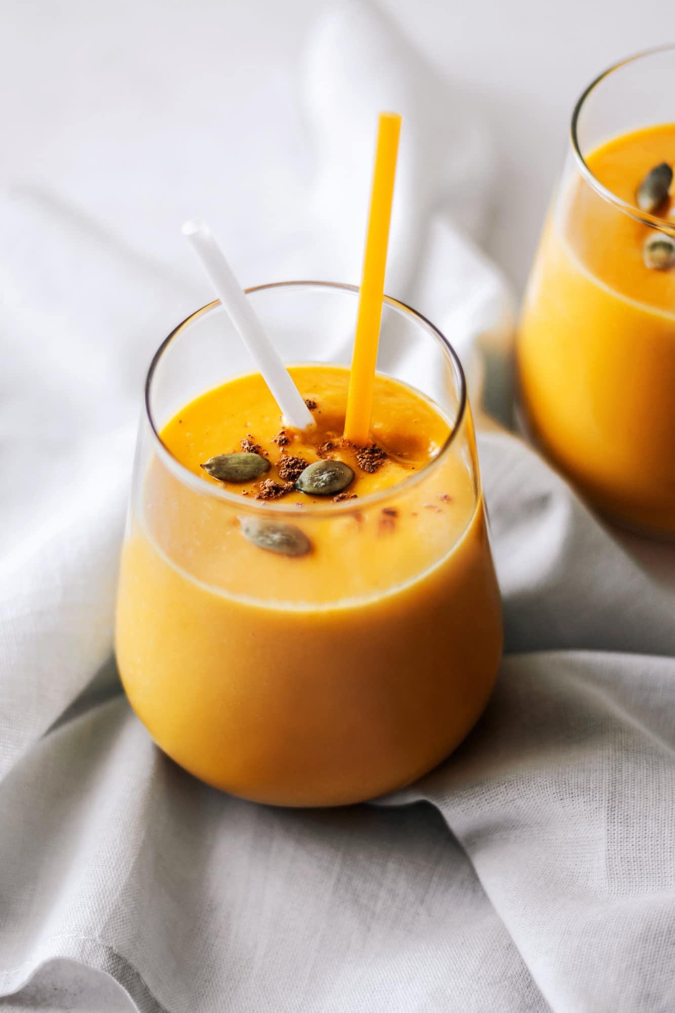 pumpkin smoothie in a glass cup with straws and pumpkin seeds sprinkled on top.