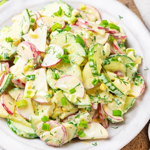 wooden-board-under-white-plate-with-radish-cucumber-salad-with-dill-and-green-onion-and-dressing-and-a-silver-spoon