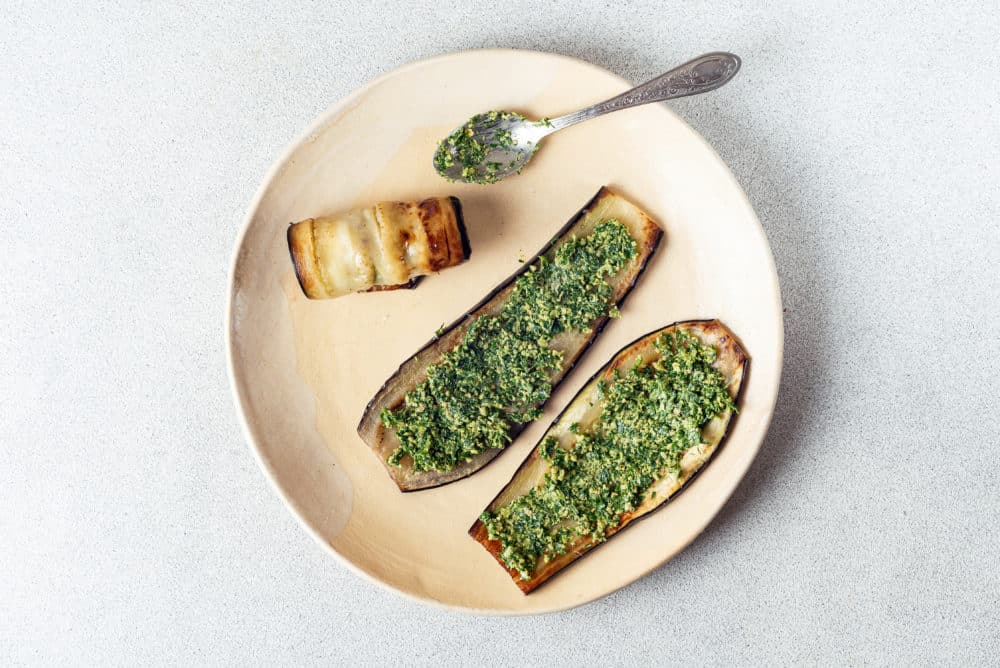 eggplant slices with herb mix spread out on them and one slice rolled up on a beige plate with a spoon.