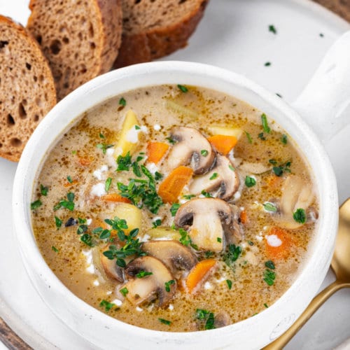 A white bowl of mushroom soup with slices of bread and a spoon on the side.