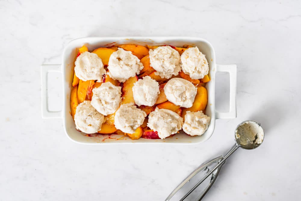 Peach cobbler ready to bake with dough pieces on top and an icecream scoop on the side.