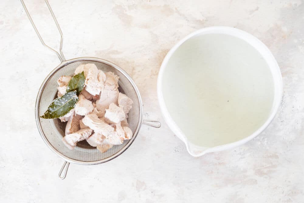 Chicken and bay leaves in a sieve with a bowl of broth on the side.