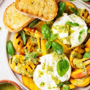 a white plate with grilled peaches burrata cheese balls fresh green basil and salad dressing.