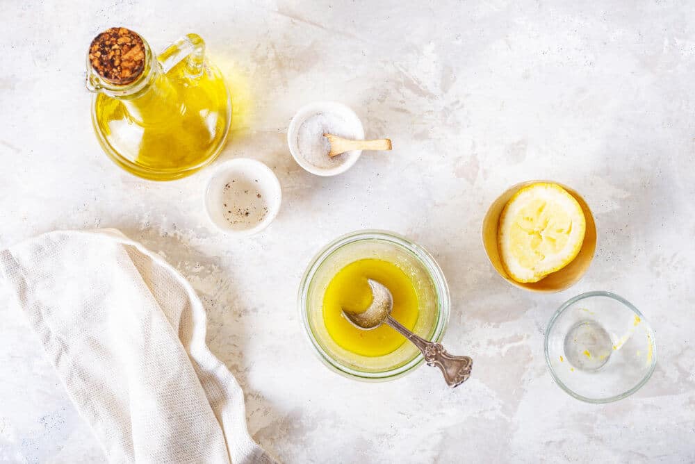 dressing in a glass bowl with empty ingredient bowls on the side and olive oil in a glass carafe and a lemon wedge.