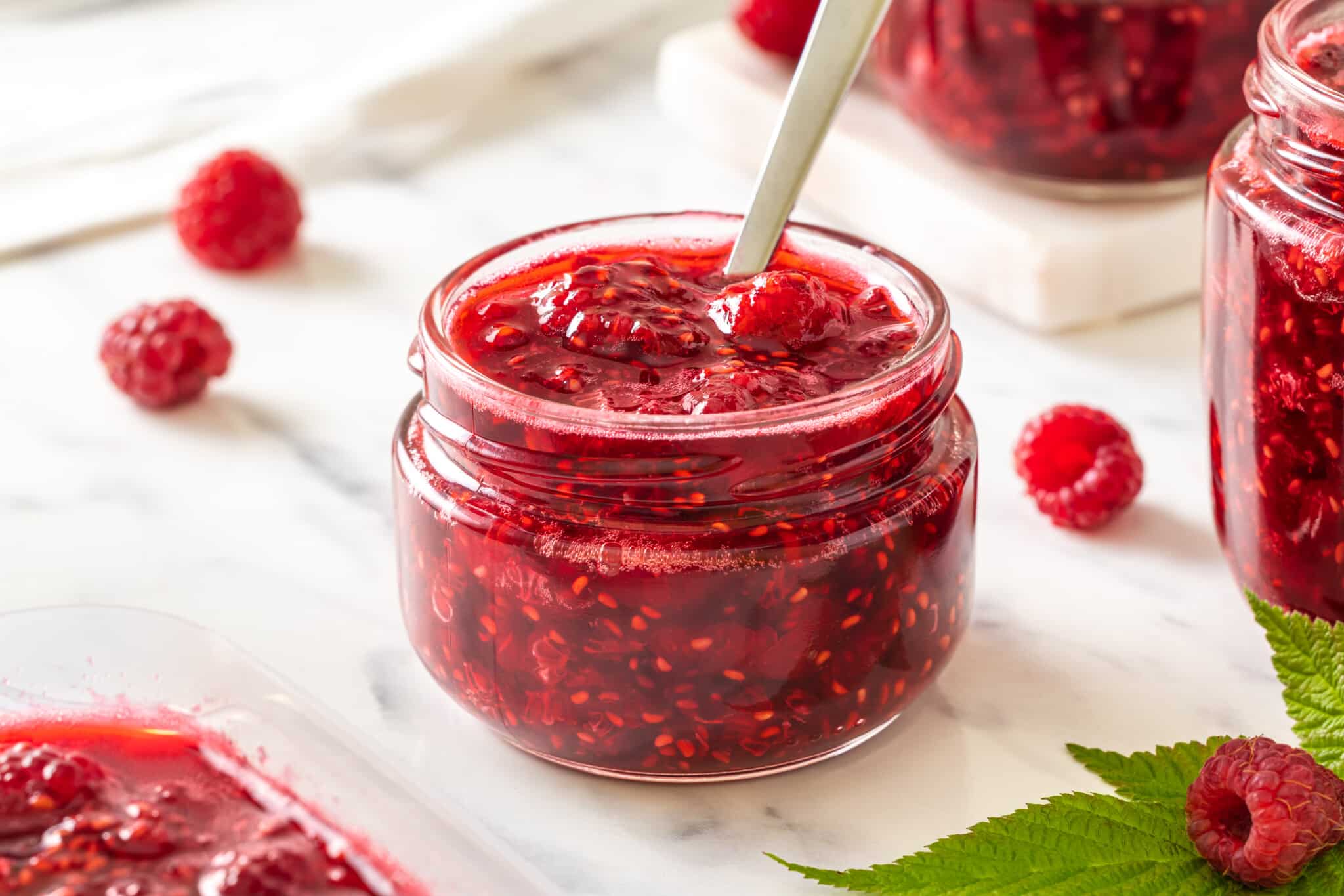 raspberry-jam-in-a-clear-jar-with-a-spoon-and-with-raspberries-and-more-jam-jars-in-the-background