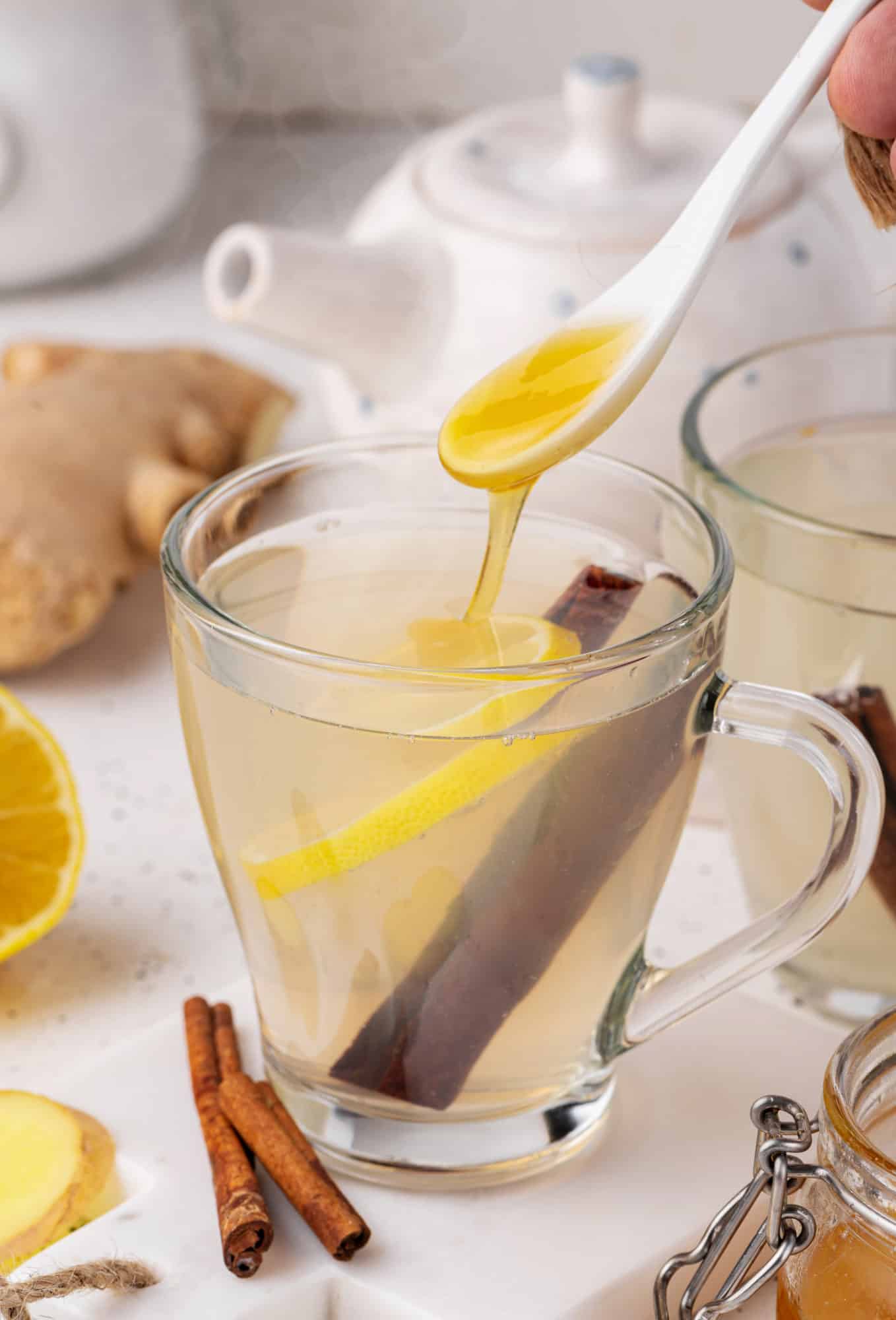 ginger tea in a glass mug with lemon and cinnamon in the tea with honey on a spoon being poured into the tea. 