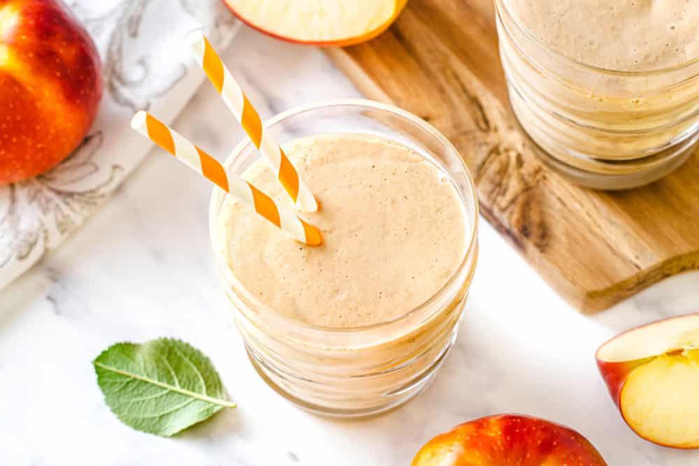 an apple smoothie in a clear glass with two straws and a wooden board on the side.