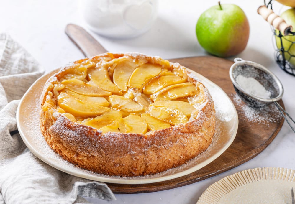 apple-cake-on-a-white-plate-on-a-wooden-board-with-a-sieve-with-powdered-sugar-in-it