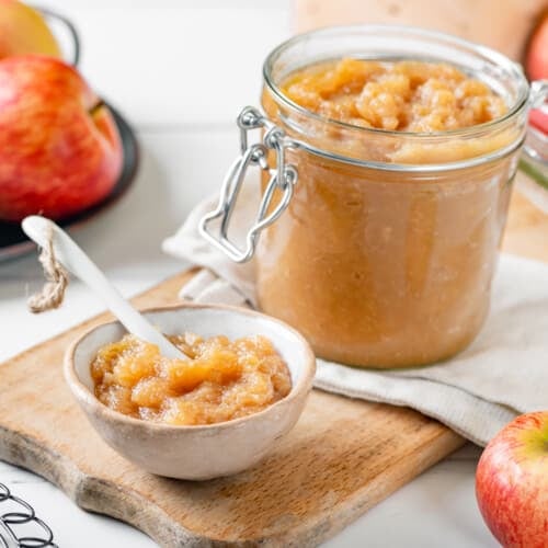 A glass jar and a white bowl of apple jam with a spoon in the bowl and apples all around.