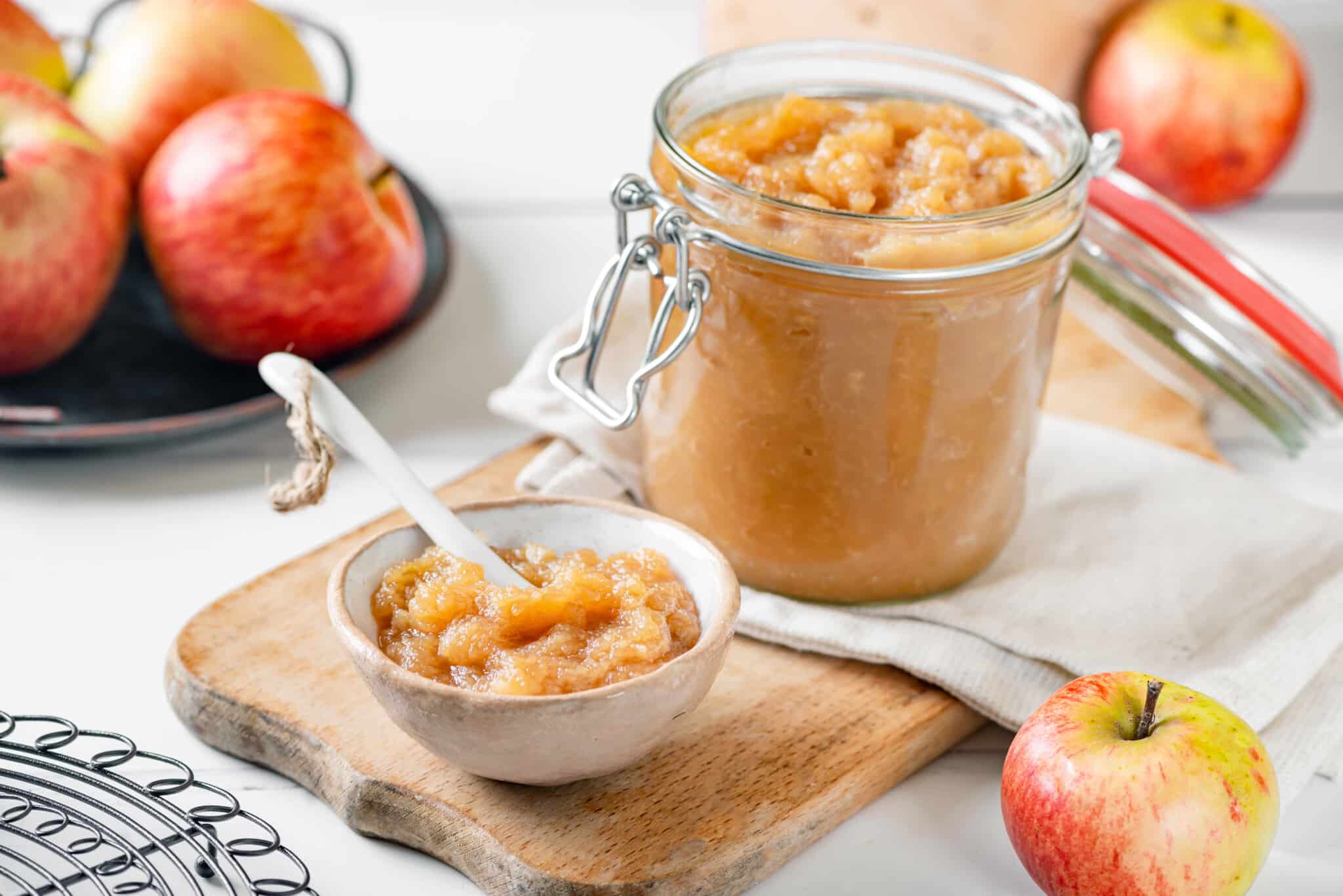 A glass jar and a white bowl of apple jam with a spoon in the bowl and apples all around.