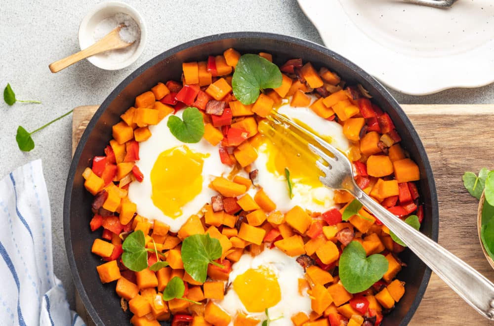 Sweet potato and eggs in a skillet with silver forks and a sprinkle of micro greens on a wooden board.