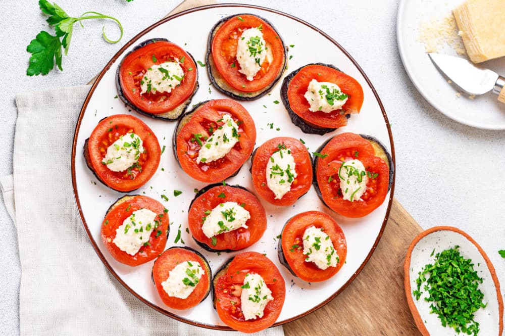 top view of tomato and eggplant slice appetizers on a white plate with a brown rim and with more chopped parsley on the side in a small white bowl.