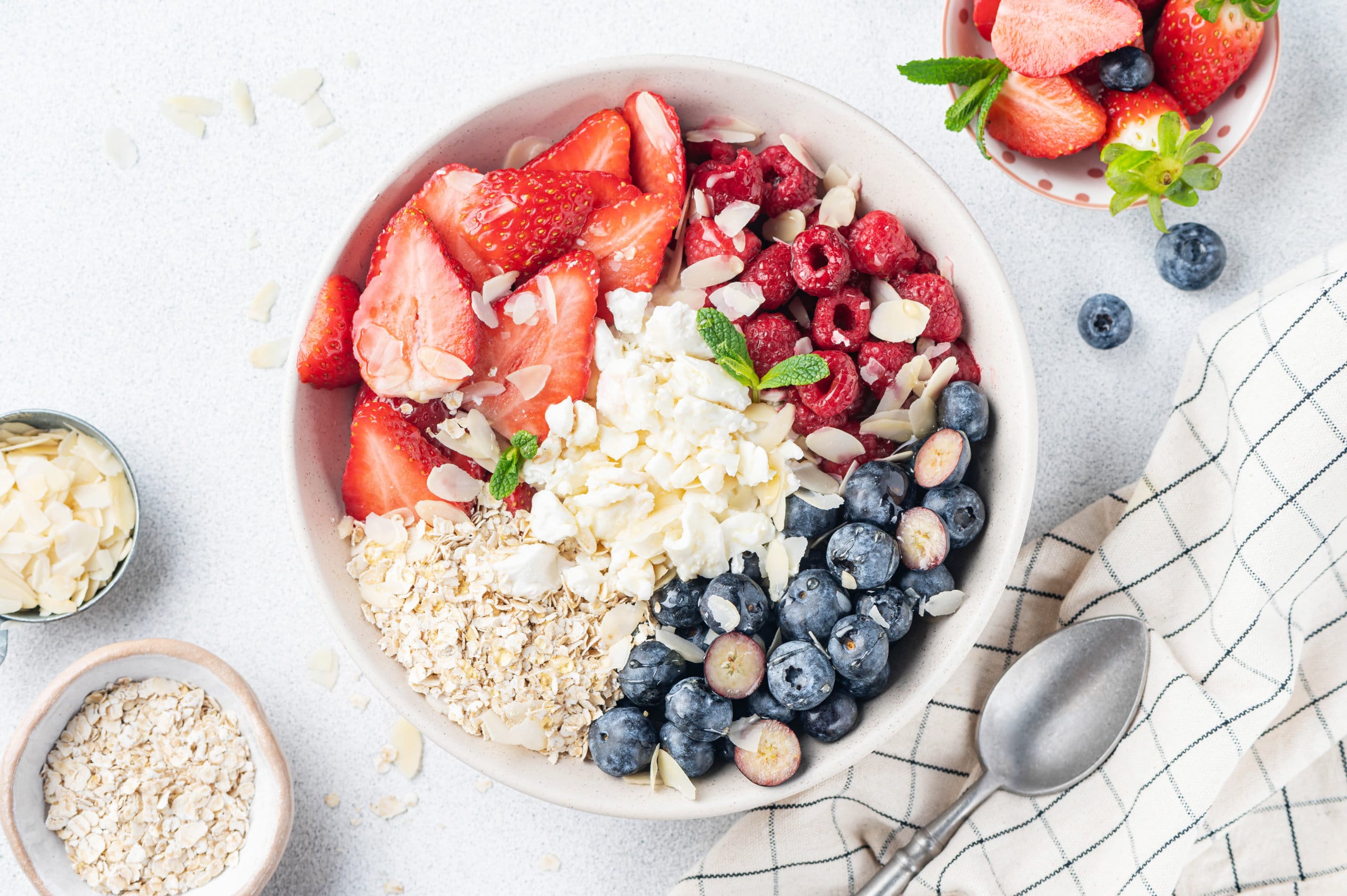 A bowl of cottage cheese and fruit with oats drizzled honey and silvered almonds and a spoon on the side.