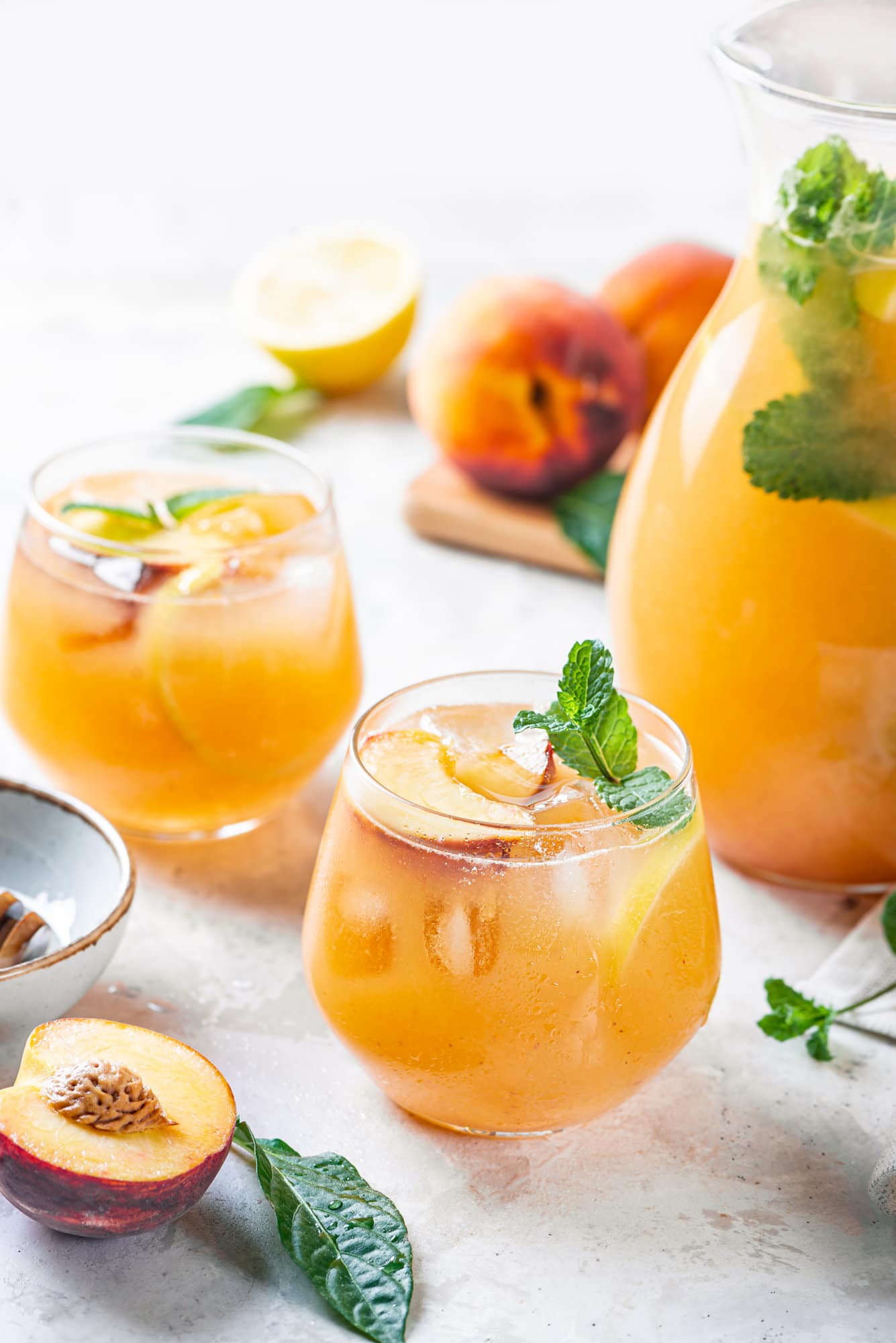 Peach lemonade in a glass cup with a pitcher on the side and fresh peaches around.