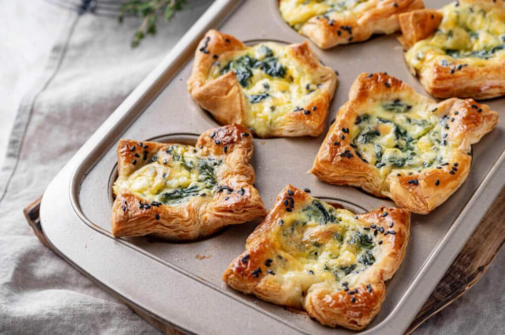 baked spinach puffs in a muffin tray on a wooden board with a towel.