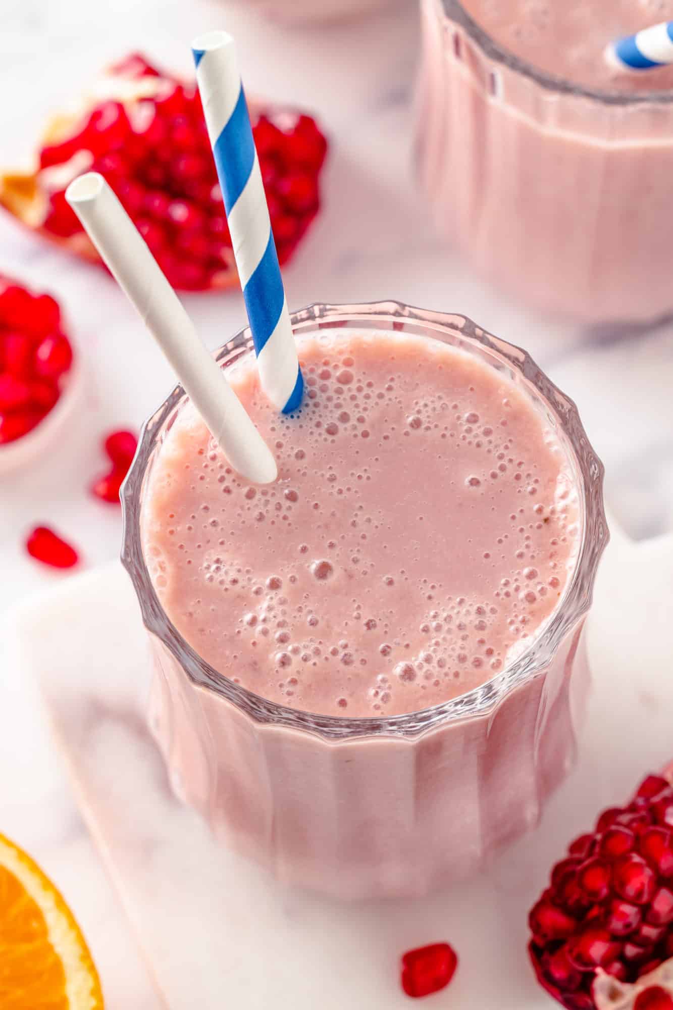 top view of a smoothie in a glass made of pomegranate with two straws in the smoothie and more ingredients on the side.