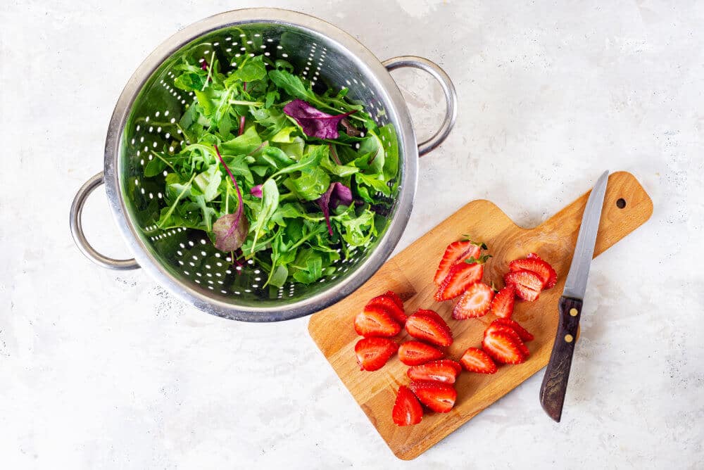 greens in a silver sieve with chopped strawberries on a wooden board with a knife.